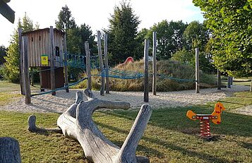 Baumstamm und Wippe vor Spielplatzhügel mit Rutsche, Kletterseilen und Baumhaus